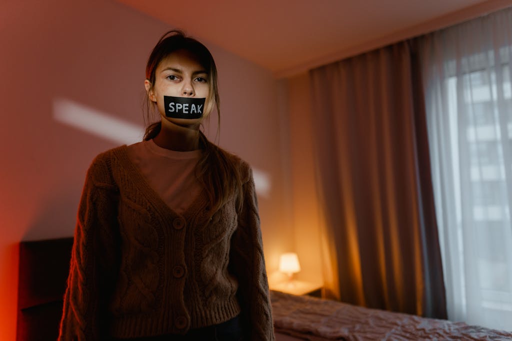 A woman stands in a dimly lit bedroom with tape reading 'Speak' over her mouth, symbolizing silence and oppression.